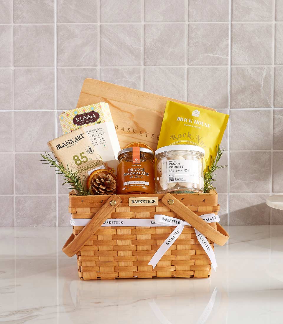 The Hearty Eats Hamper features a wicker gift basket brimming with chocolate, orange marmalade, vegan cookies, and snacks. Adorned with decorative rosemary and a pinecone, this delightful ensemble graces the kitchen countertop.