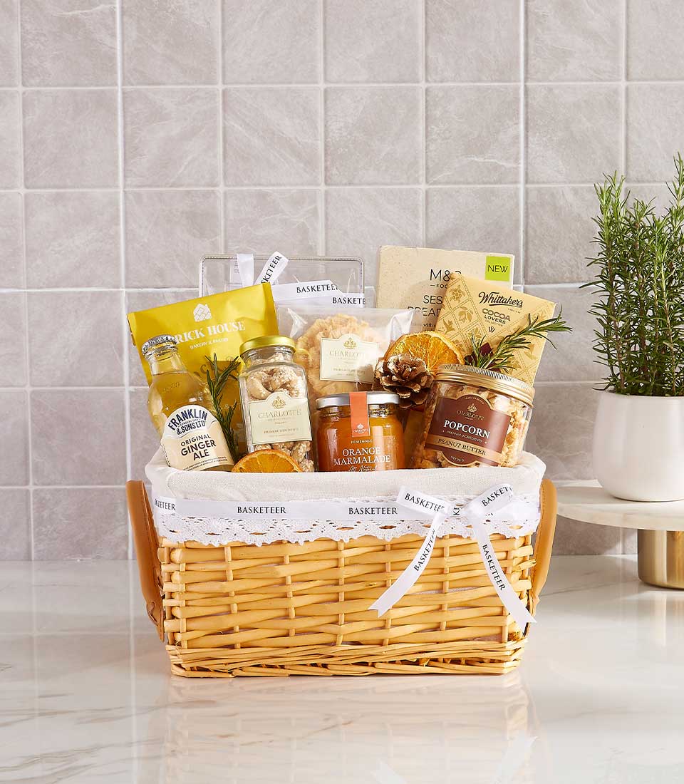 A Share the Love Gift Basket brimming with gourmet snacks graces the marble countertop. It includes popcorn, jam, tea, and biscuits, all adorned with lace trim and ribbons. Nearby, a small potted plant adds charm against the tiled backsplash.