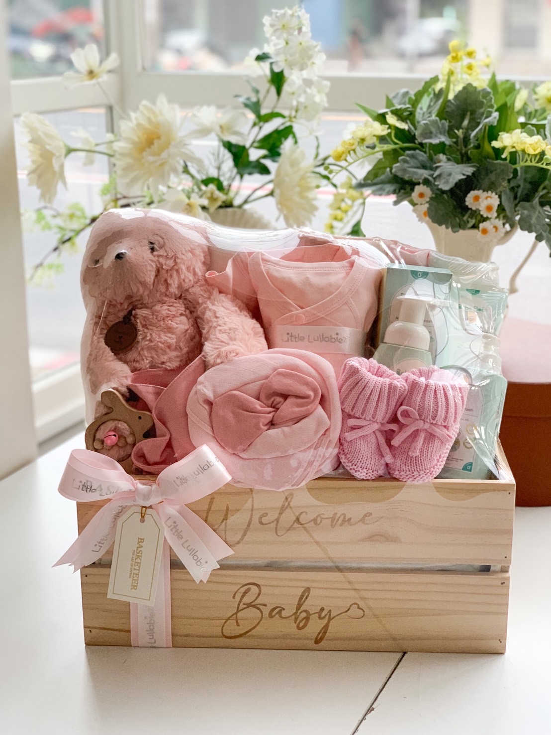 Pink baby gift box with teddy bear, romper, knitted booties, baby bottle, and care essentials in a wooden crate.