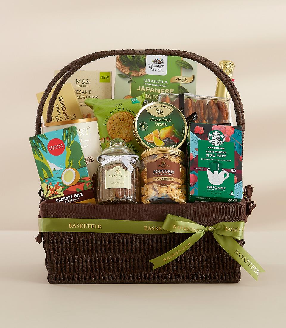 A brown wicker basket filled with a variety of gourmet treats, including granola, Japanese snacks, chocolate, and a bottle of champagne.