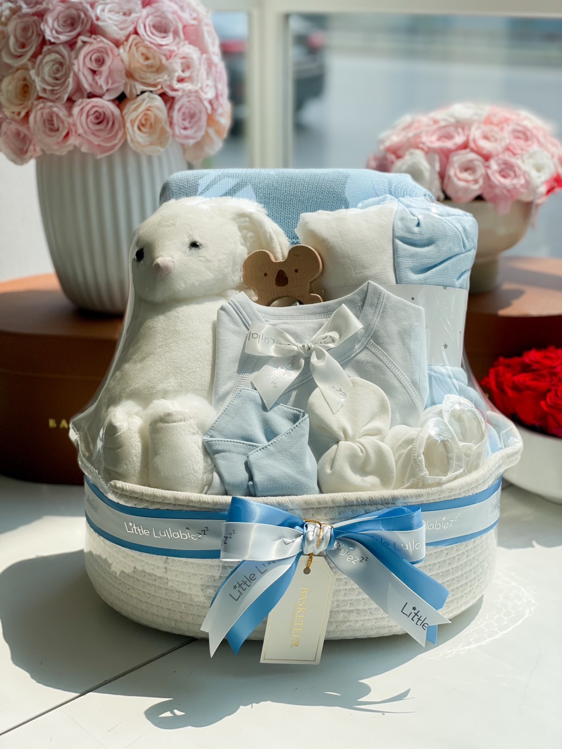 A beautifully arranged baby boy gift basket in blue, featuring soft baby clothes, a plush bunny, a baby blanket, a wooden teether, and newborn essentials, wrapped with a blue ribbon from Basketeer.