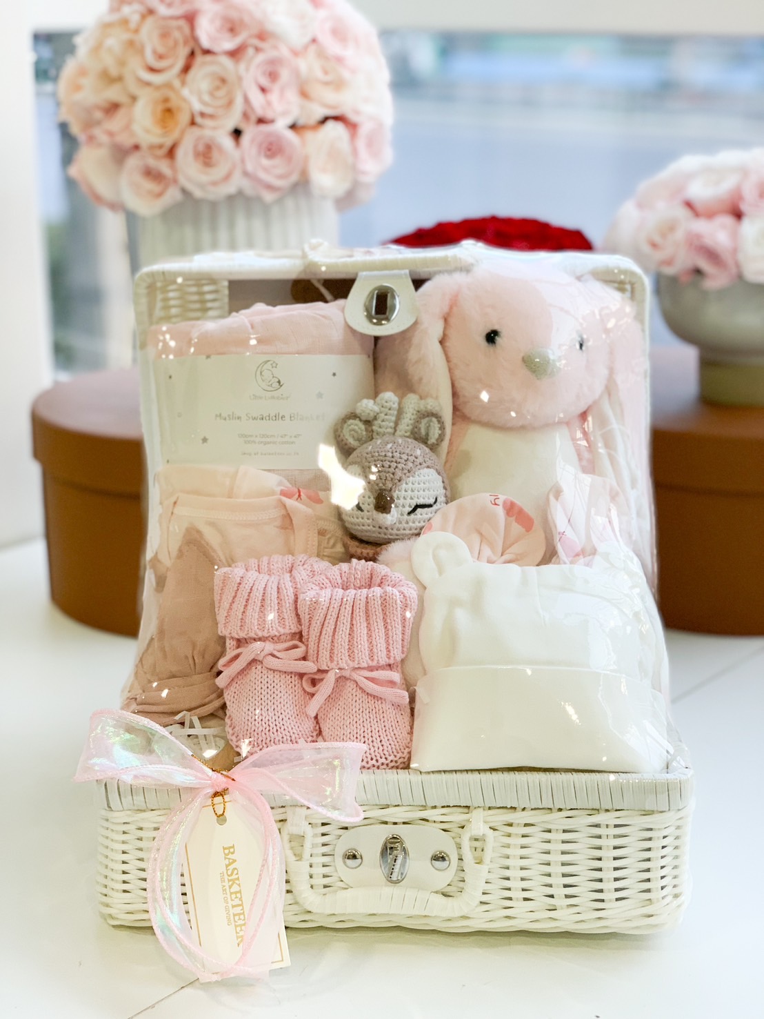 A charming baby girl gift basket in soft pink, featuring a plush bunny, knitted booties, a swaddle blanket, a crochet rattle, and baby essentials in a white wicker basket with a pink ribbon.