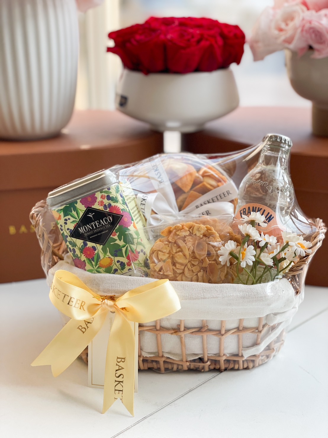 A beautifully curated gourmet gift basket featuring Monteaco premium tea, crispy almond cookies, handcrafted pastries, and a refreshing Franklin soda. Wrapped in a woven basket with a golden ribbon and floral decorations, perfect for any occasion.