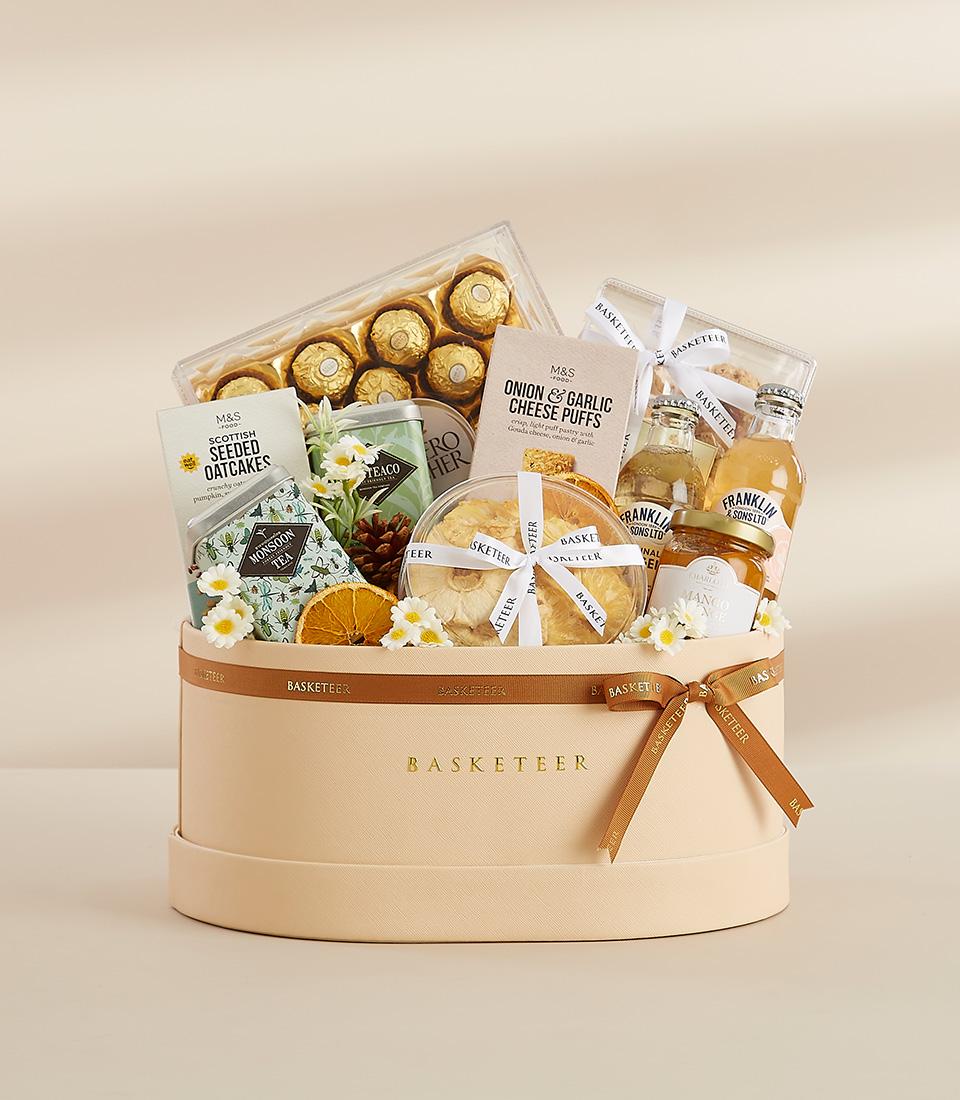 A beige gift box filled with a variety of gourmet treats, including chocolates, snacks, tea, and flowers.