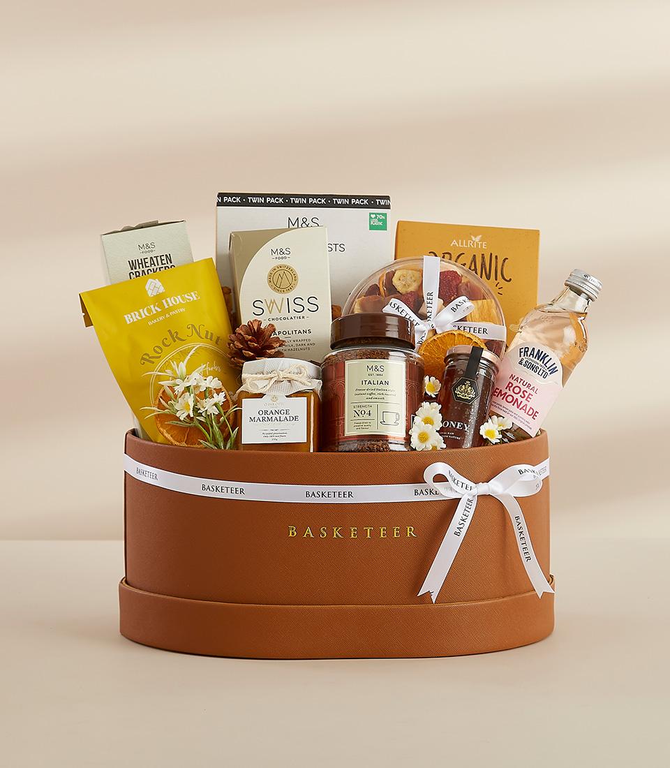 A round gift box filled with a variety of gourmet treats, including chocolates, snacks, tea, and flowers.