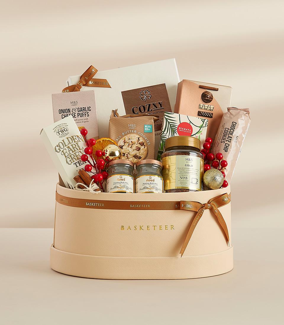 A round gift box filled with a variety of gourmet treats, including chocolates, snacks, tea, and festive decorations.