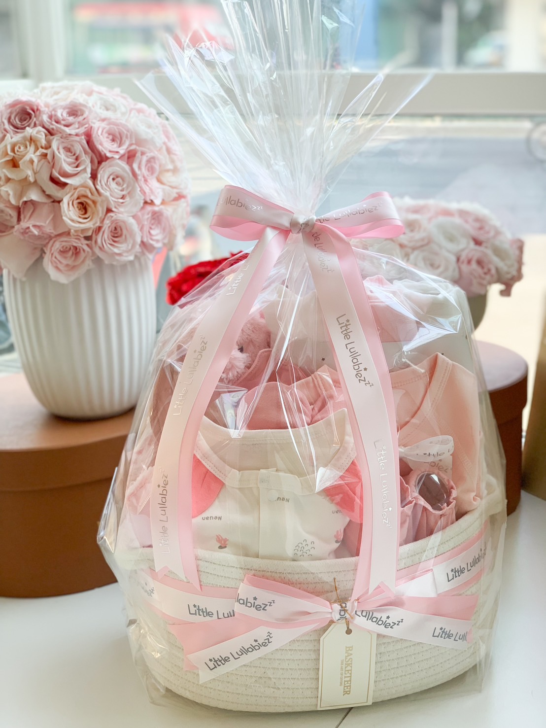 A beautifully curated baby girl gift basket in soft pink, featuring adorable newborn essentials like baby clothing, plush toys, blankets, mittens, and more. Thoughtfully arranged in an elegant fabric basket with a delicate pink ribbon—the perfect gift for new moms and baby showers.