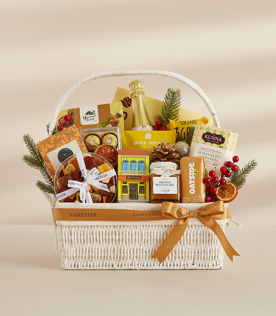 A white wicker basket filled with a variety of gourmet treats, including chocolates, snacks, tea, and festive decorations.