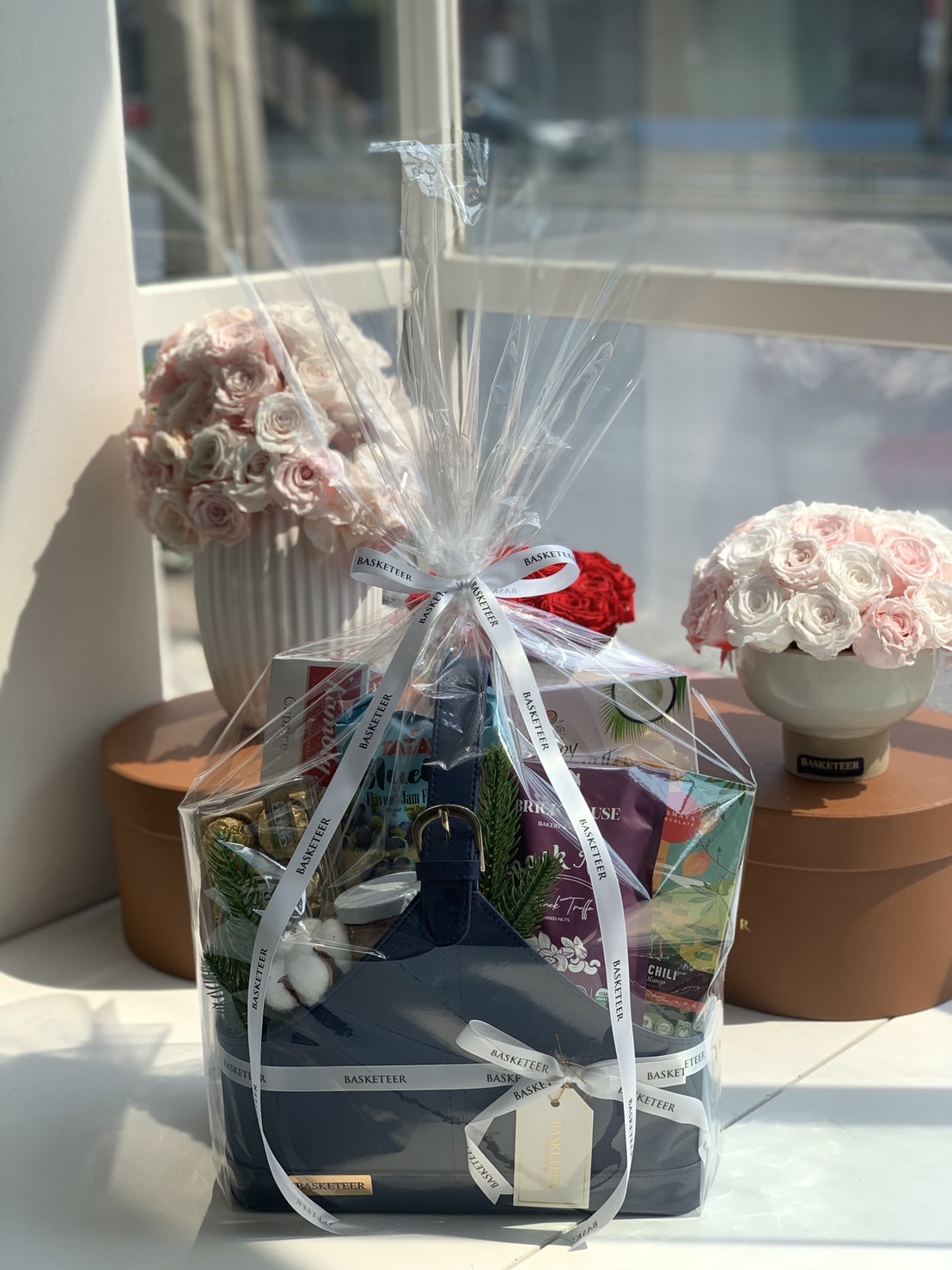 A luxury gourmet gift basket wrapped in clear cellophane with white Basketeer ribbons, featuring a navy blue leather tote filled with premium treats.