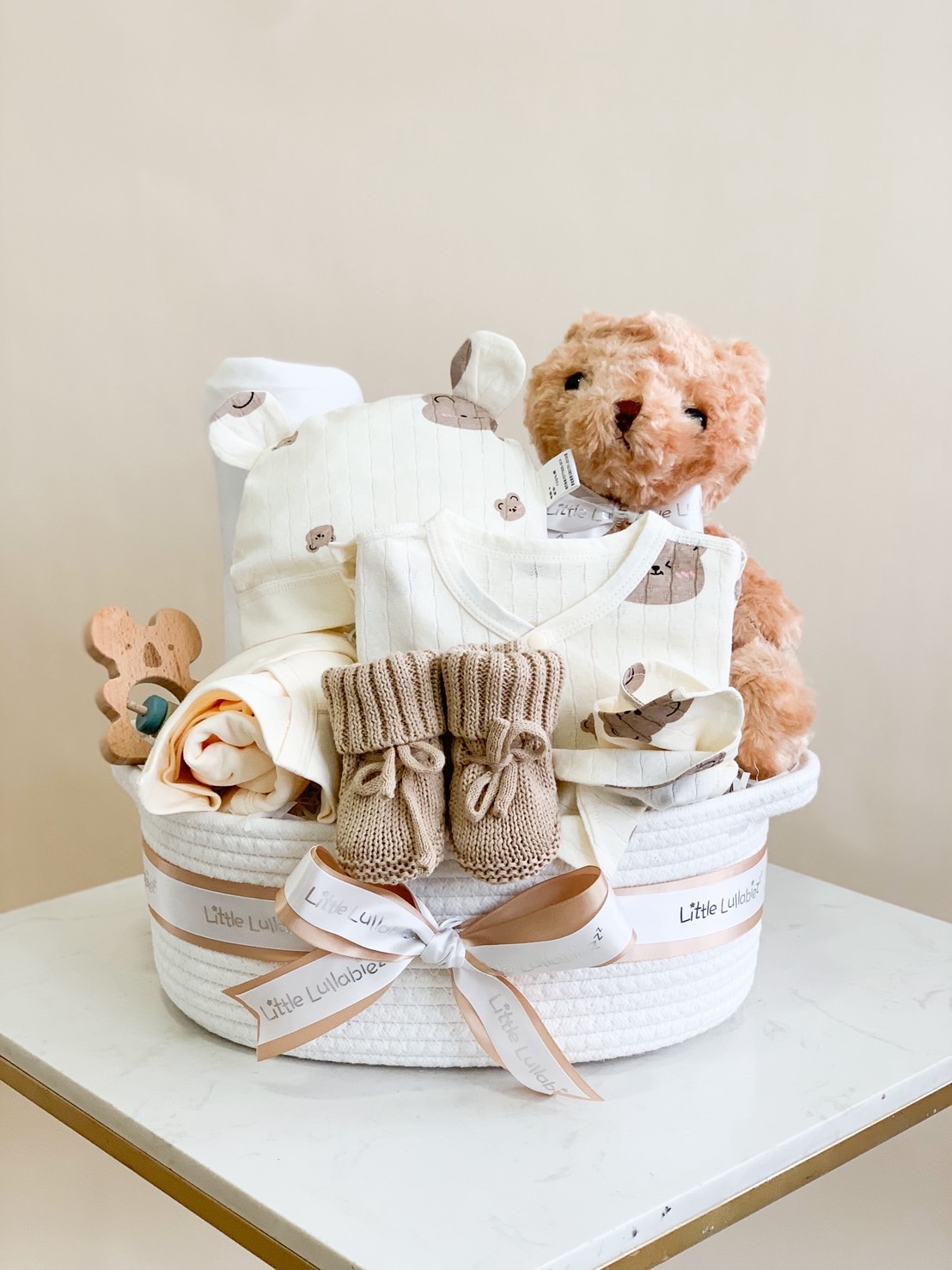 Neutral-toned baby gift basket featuring baby clothes, beige knit booties, teddy bear, and essentials in a white woven basket.