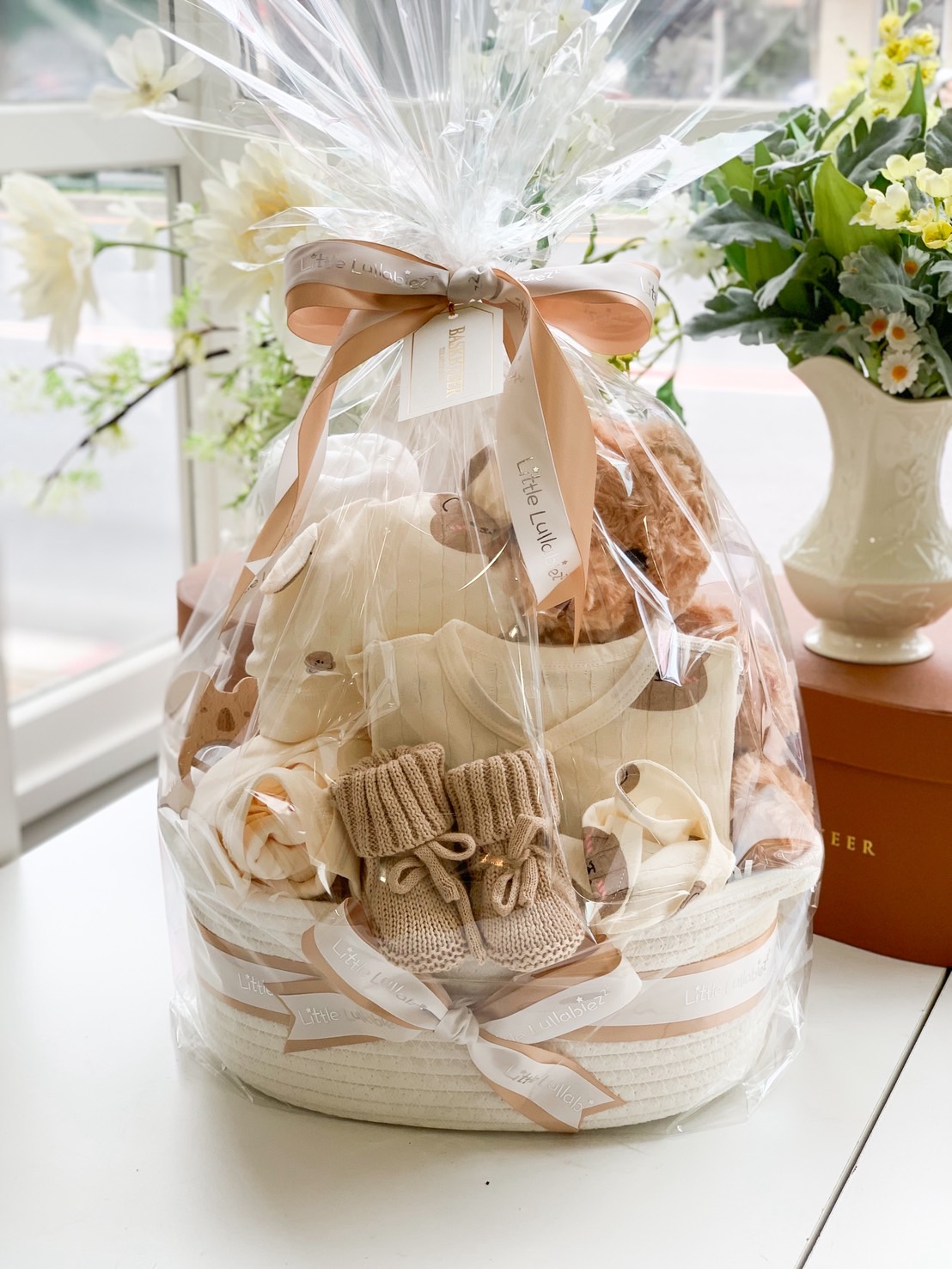 Wrapped baby gift basket with beige-toned babywear, knit booties, teddy bear, and elegant ribbon in clear gift wrapping.