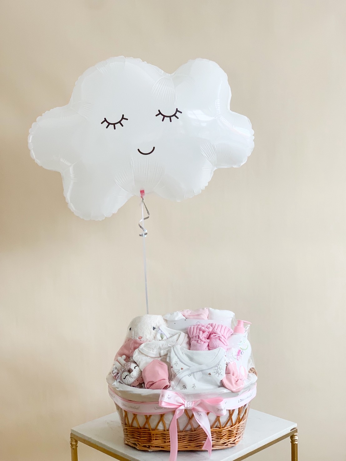 Baby girl gift basket with pink and white clothing, plush toy, baby essentials, and a smiling white cloud balloon, tied with a pink ribbon.