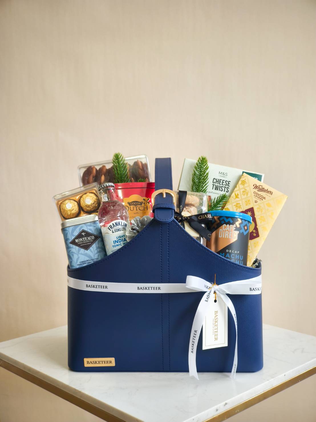 Navy leather gift basket filled with chocolate, cookies, nuts, coffee, and premium snacks with a white ribbon