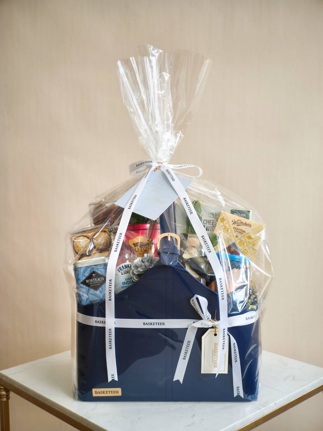 Gift-wrapped navy leather basket filled with chocolates, snacks, and premium treats, sealed in clear plastic wrap with white ribbons