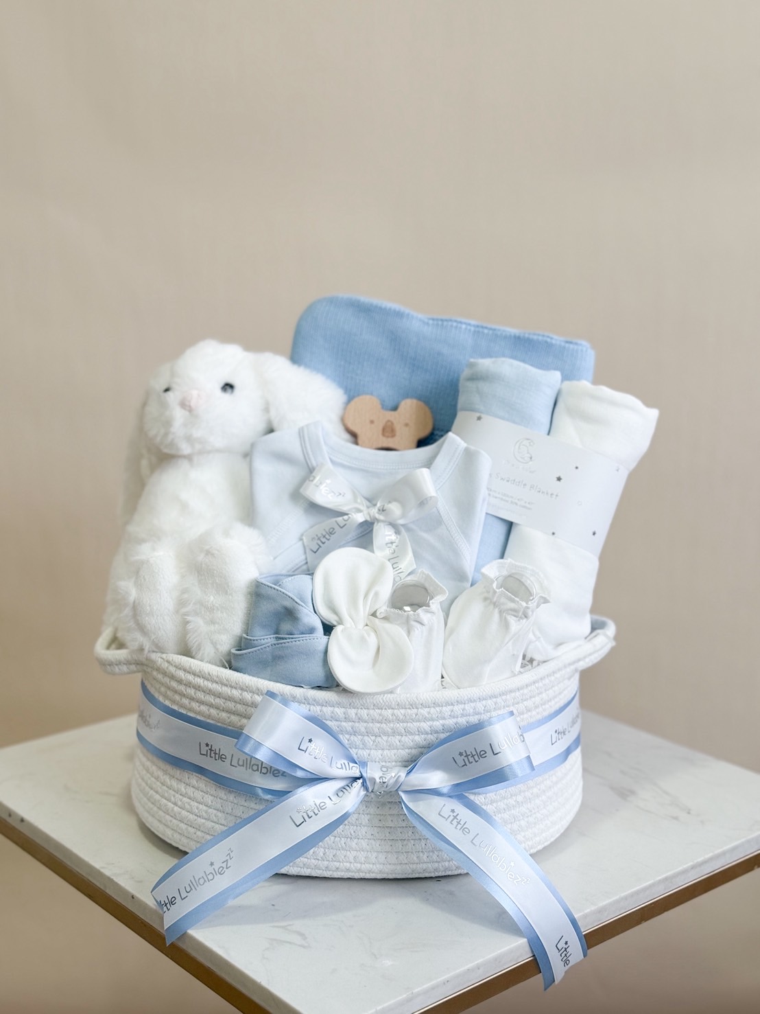 White and blue baby boy gift basket featuring soft bunny plush, baby clothes, swaddle blanket, and accessories.