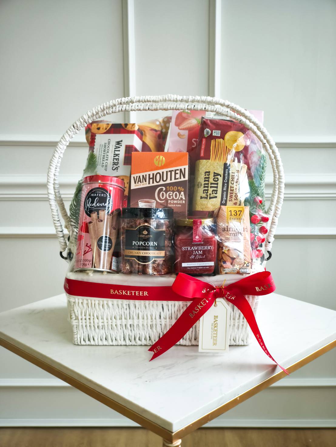 Premium white wicker gift basket with imported snacks, cocoa powder, biscuits, jam, popcorn, and health drinks, tied with a red satin ribbon.
