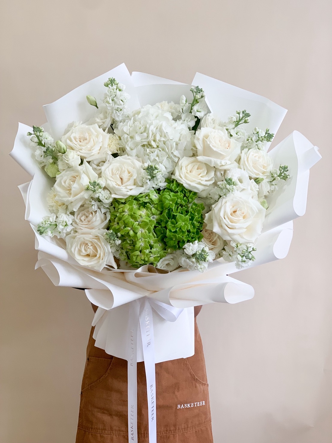 A large hand-held bouquet of white roses, hydrangeas, and green florals wrapped in white paper with a satin ribbon.