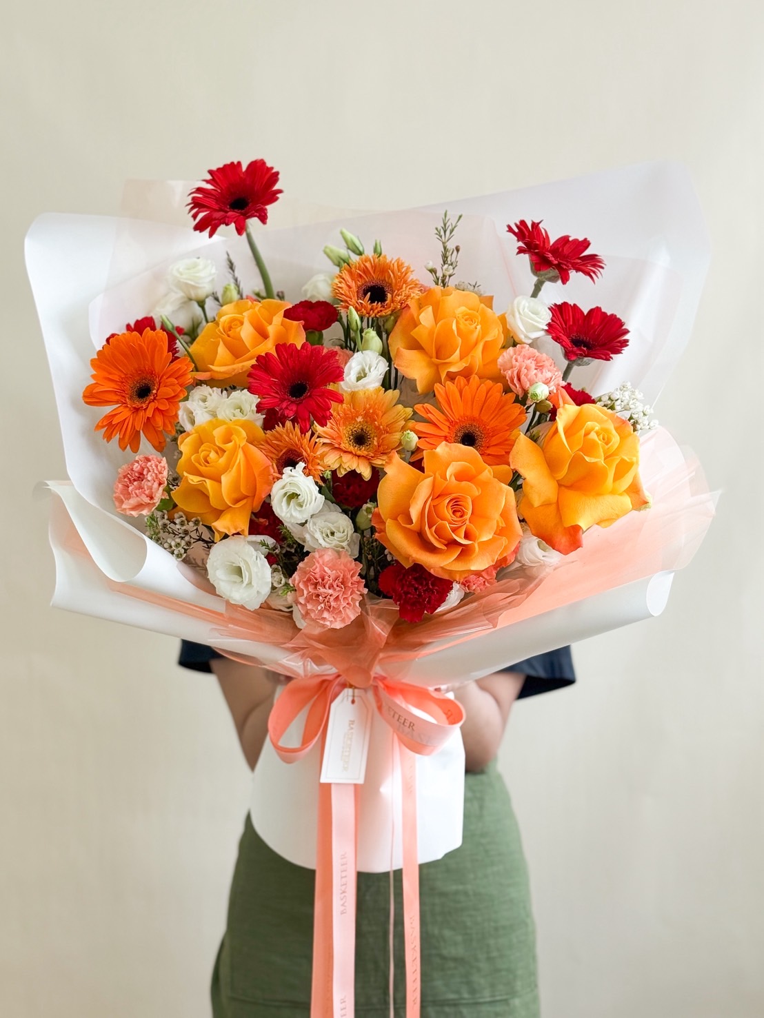 Cheerful bouquet with orange roses, red gerberas, white lisianthus, and pink carnations, wrapped in white and peach paper with satin ribbon.