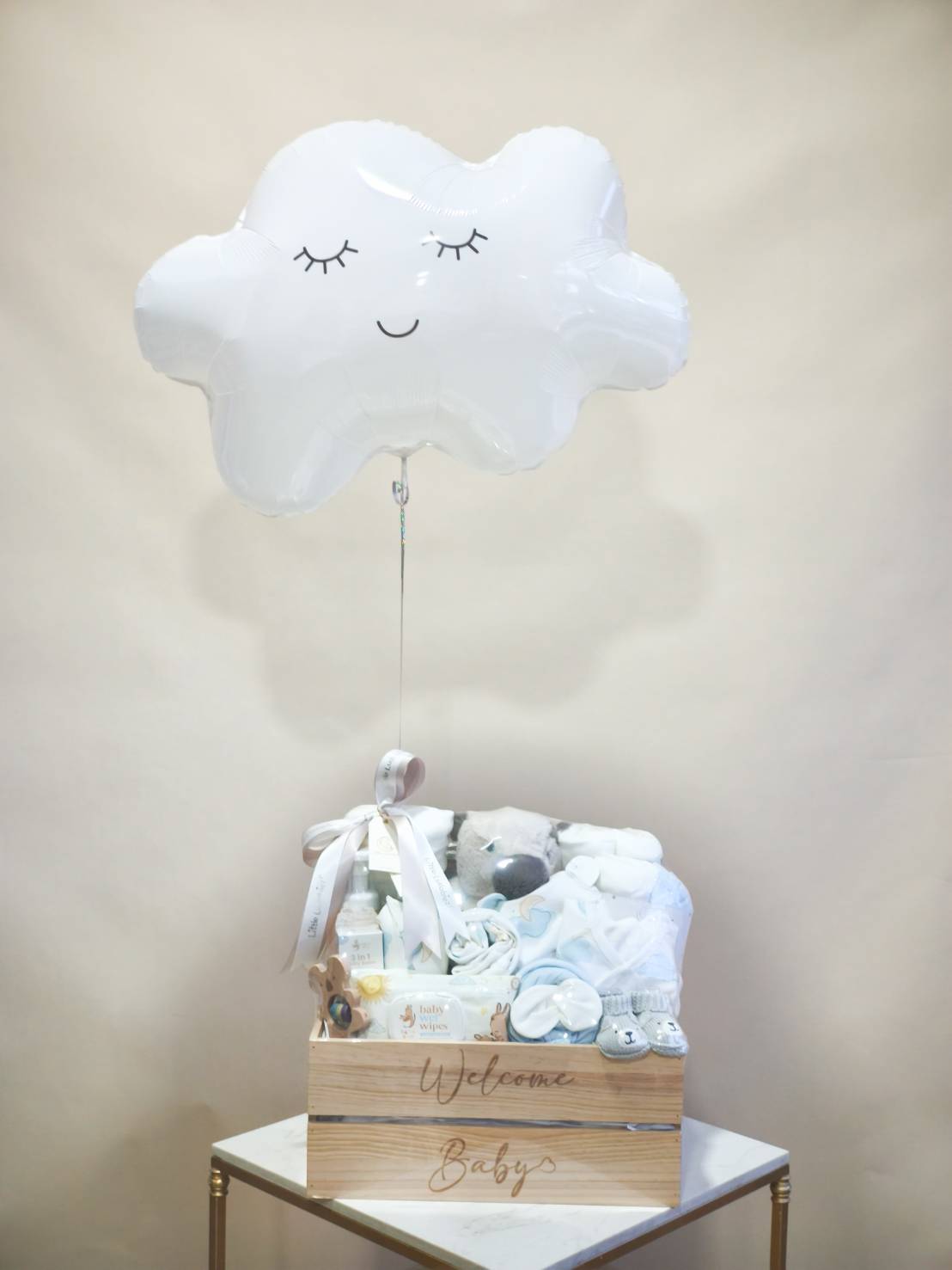 Newborn wooden gift box with baby clothes, socks, wipes, plush toy, and essentials, decorated with a white cloud balloon and satin ribbon.