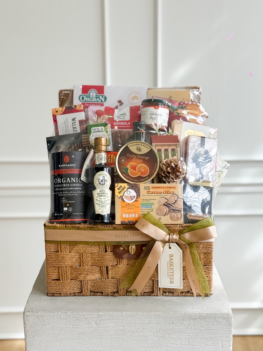 A Healthy Gourmet Gift Hamper filled with assorted gourmet food items, including snacks, olive oil, and biscuits, wrapped with a gold ribbon and tag, displayed on a white pedestal against a light background.