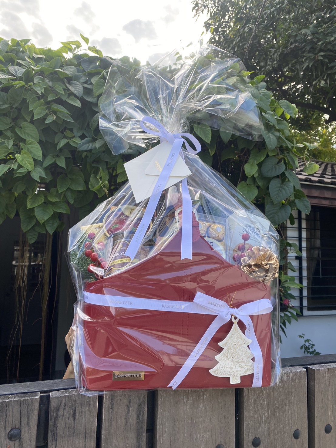 A festive red gift basket wrapped in clear plastic and tied with an elegant white ribbon, featuring a Christmas tree tag and filled with premium treats, placed outdoors with lush greenery in the background.