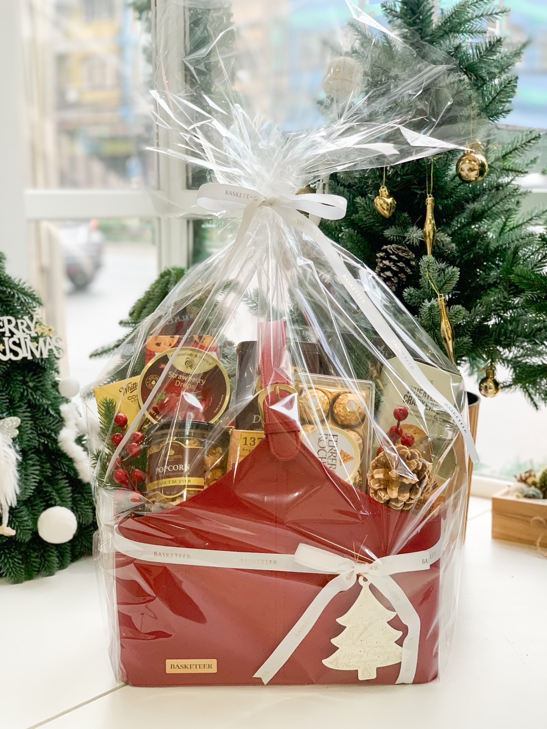 A festive red gift basket wrapped in clear plastic and tied with an elegant white ribbon, featuring a Christmas tree tag and filled with premium treats, placed outdoors with lush greenery in the background.