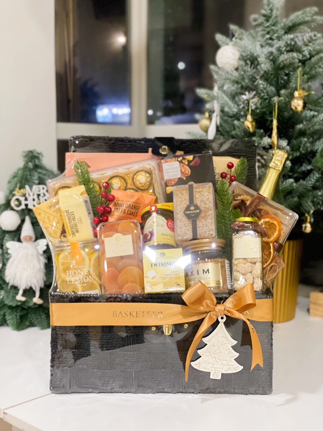 A luxurious Christmas gift hamper by Basketeer, featuring premium treats such as tea, honey lemon, dried fruits, Ferrero Rocher, and gourmet snacks, beautifully arranged in a black basket with a golden ribbon and a festive tree ornament, placed in a cozy holiday setting with a decorated Christmas tree in the background.
