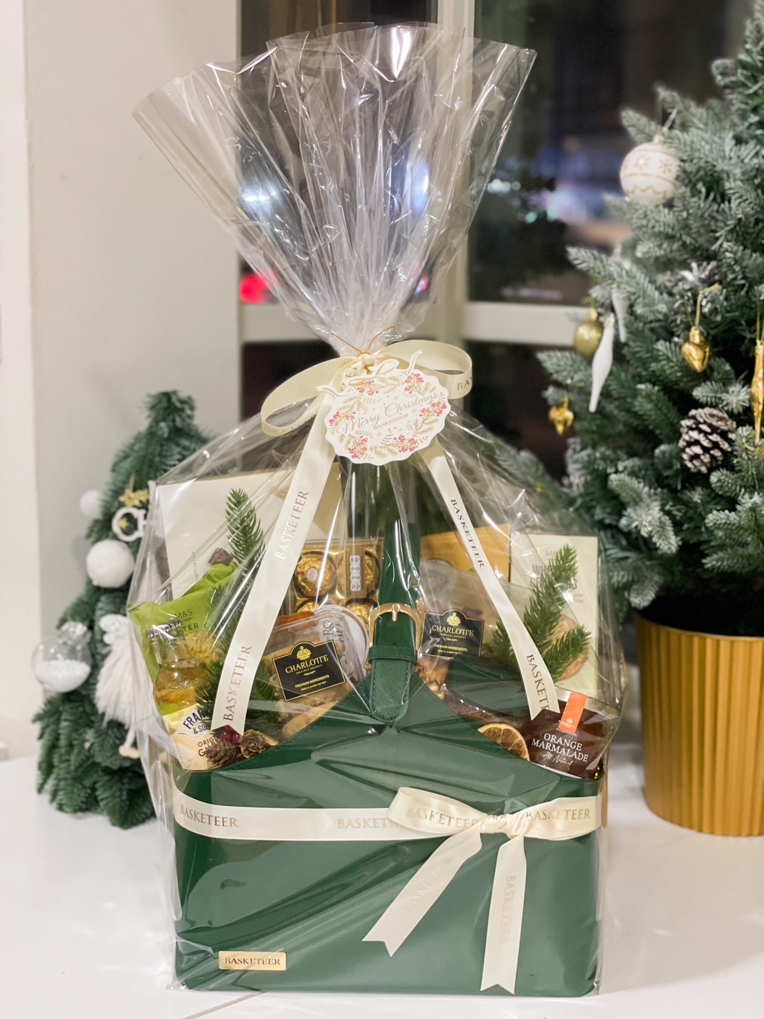 A premium Christmas gift basket by Basketeer, elegantly wrapped in clear packaging with ivory ribbons, featuring a green leather basket filled with gourmet treats, nuts, marmalade, and festive decorations, placed in a cozy holiday setting with a decorated Christmas tree in the background.