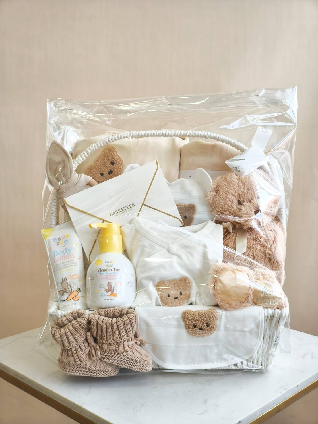 Luxury baby gift basket with cream teddy bear newborn clothes, knitted booties, plush toy, and Babi Mild baby care products, wrapped with ribbon.