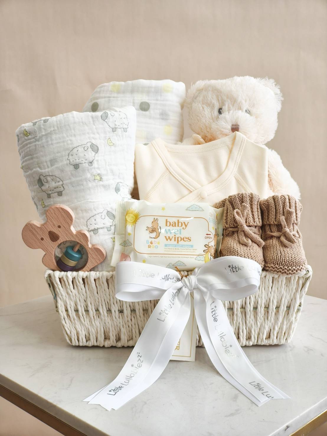 Luxury baby gift basket with cream newborn clothes, baby wipes, teddy bear, knitted booties, baby blanket, and wooden toy, decorated with ribbon.