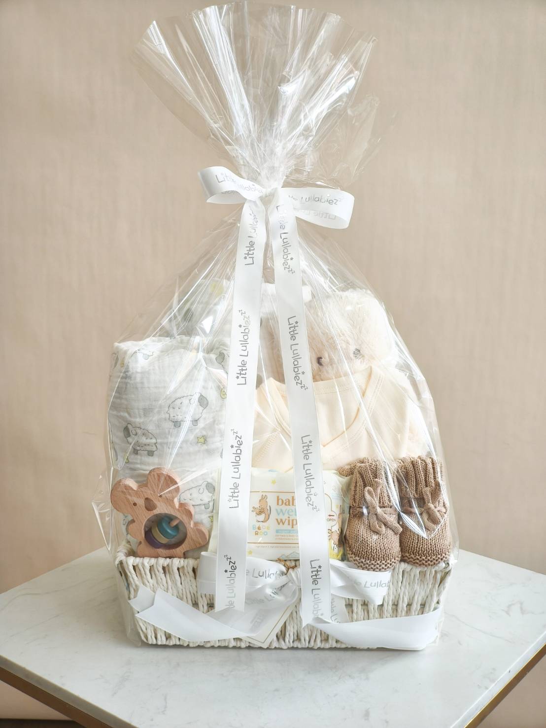 Luxury baby gift basket with cream newborn clothes, teddy bear, knitted booties, blanket, wooden toy, and baby wipes, wrapped in clear sheet.