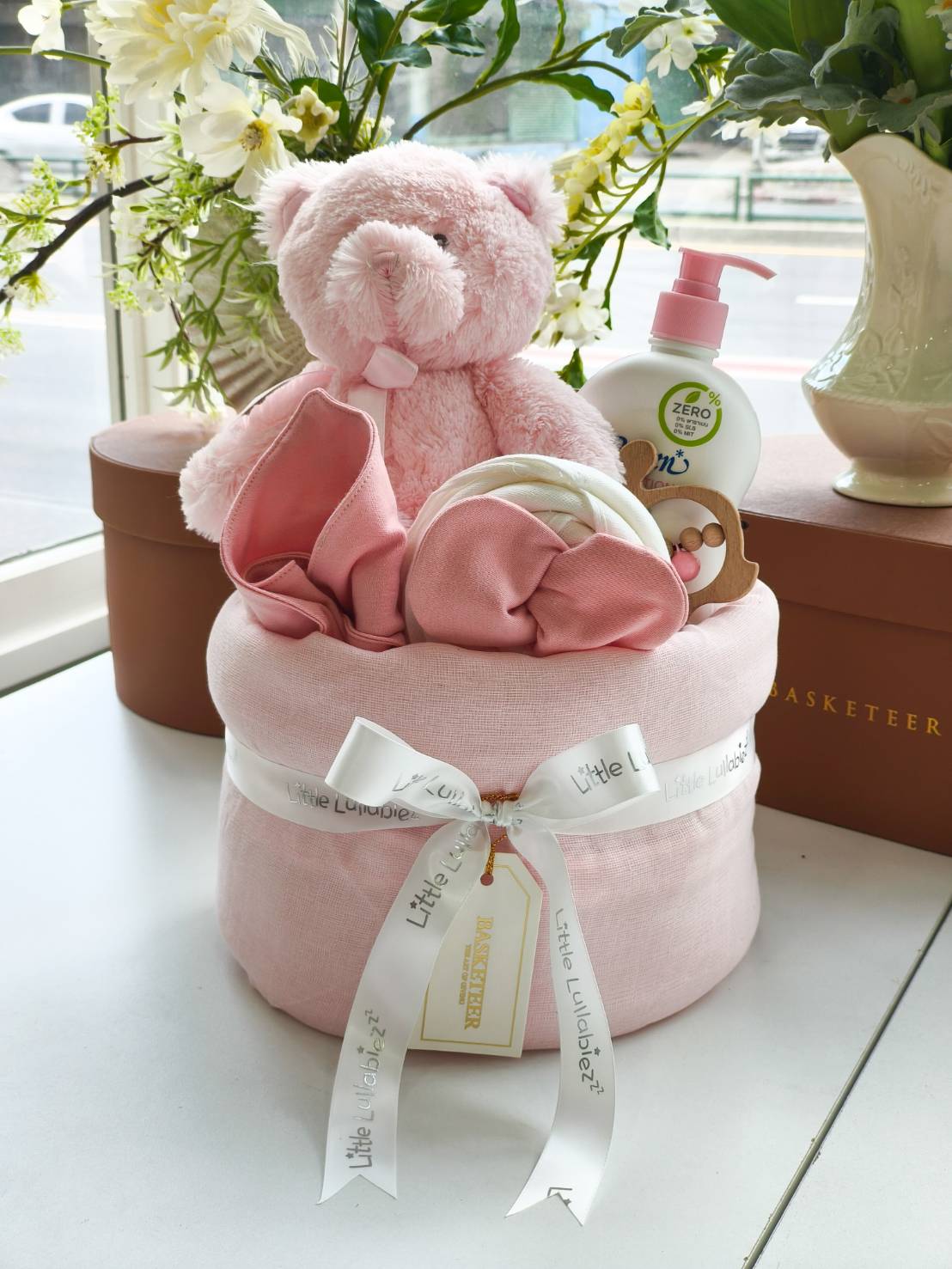 Pink baby gift basket with teddy bear, baby blanket, Zero% baby lotion, and wooden toy, decorated with Little Lullaby ribbon.