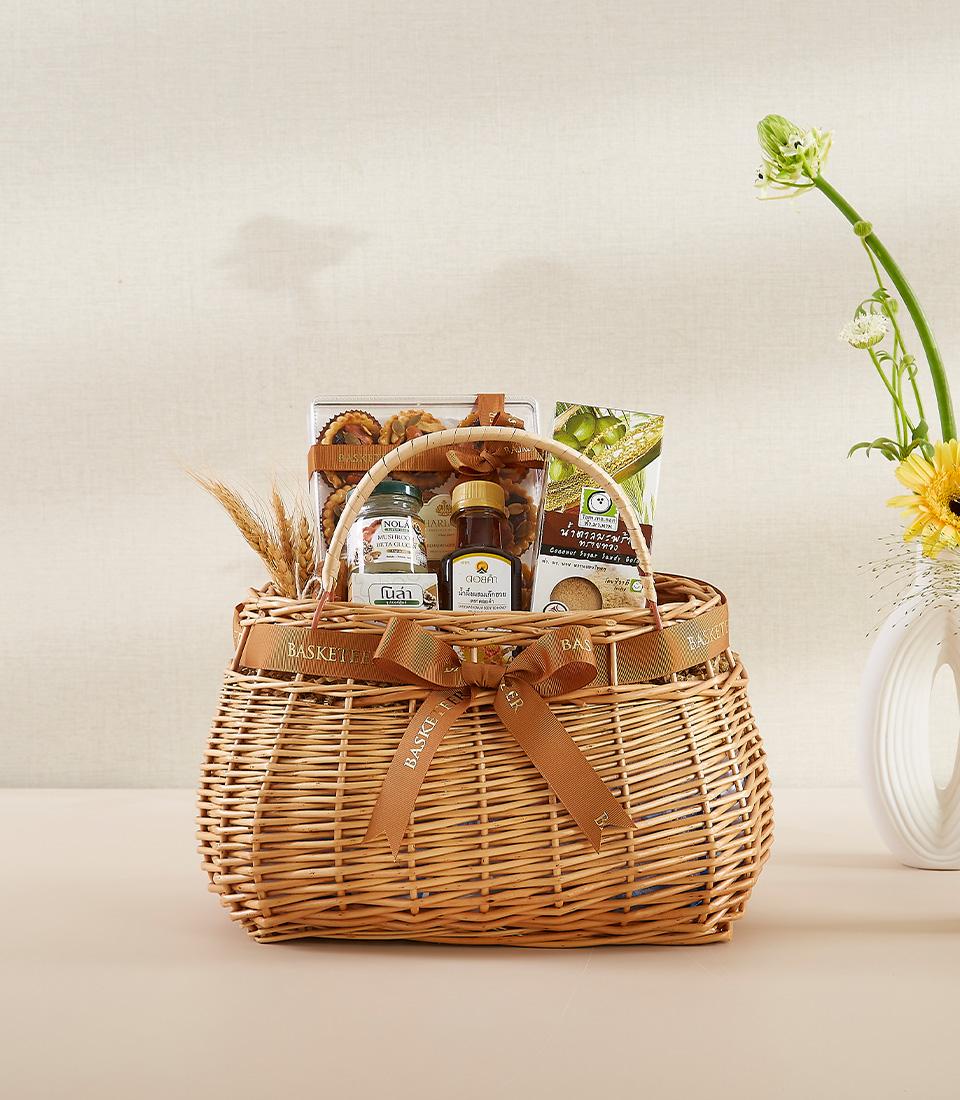 Woven basket containing olive oil, nut mix, organic rice crisps, and other gourmet items, decorated with a brown ribbon.