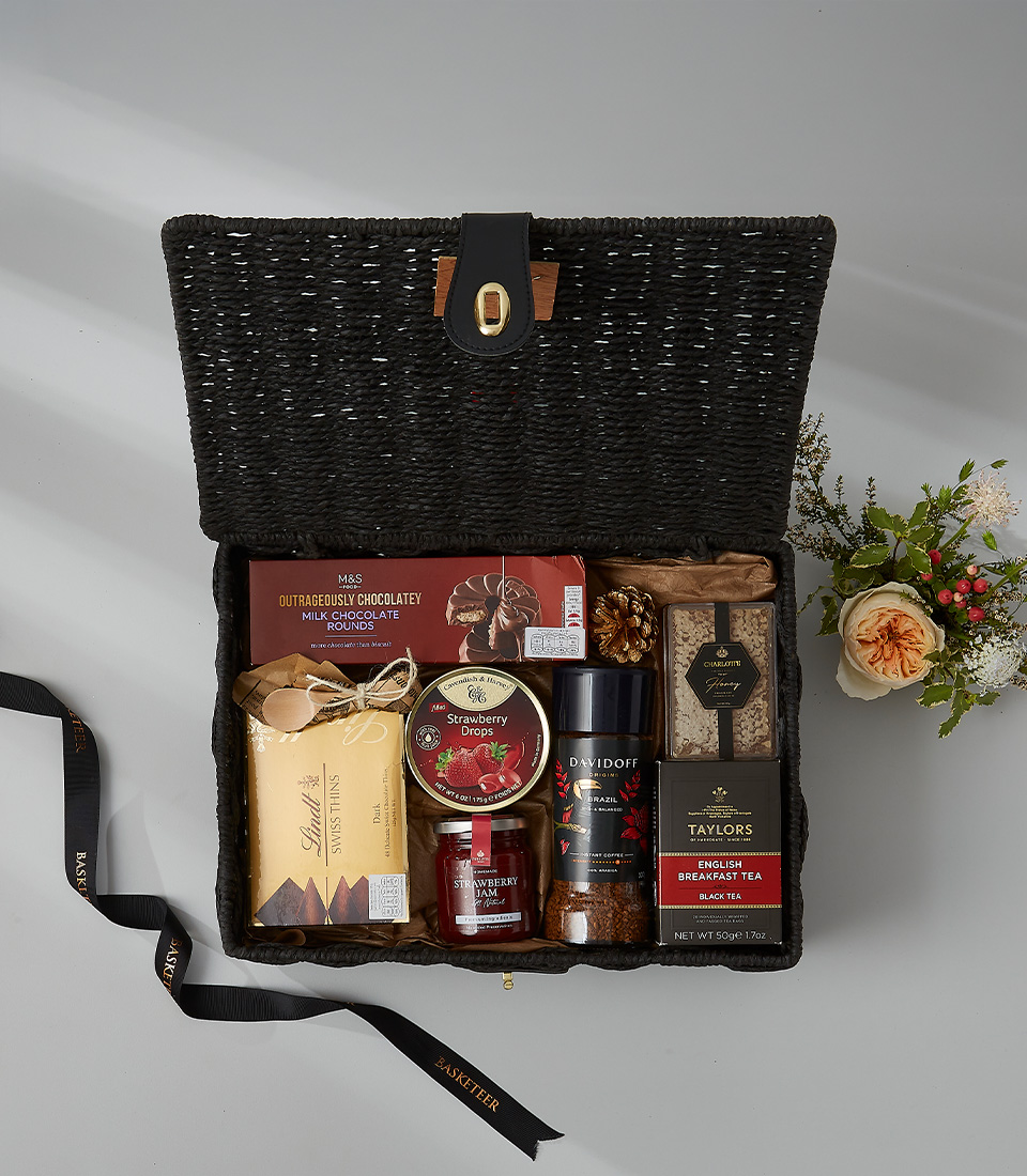 Black wicker gift hamper with tea, gourmet coffee, chocolates, strawberry jam, and sweet treats.