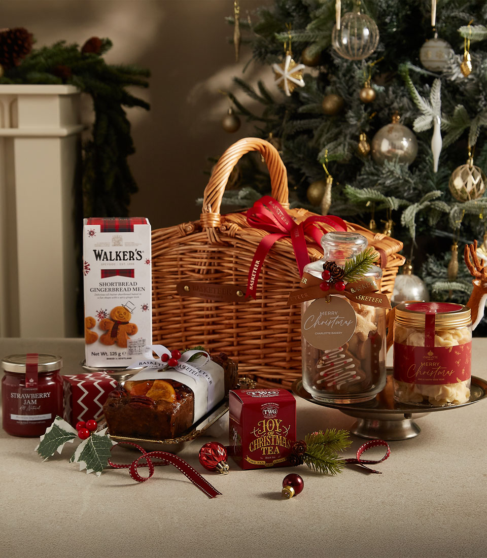 The Christmas Joy Gift Hamper includes a charming wicker basket filled with jars of cookies and jam, chocolate, and a tea canister. Set on a wooden table, it sits against the backdrop of a decorated Christmas tree with festive ornaments and greenery.
