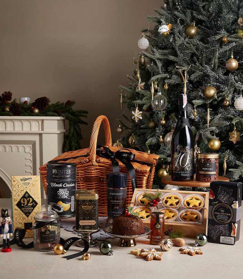 Gourmet Christmas hamper with chocolate-coated chips, coffee, Christmas pudding, and festive treats displayed near a Christmas tree.