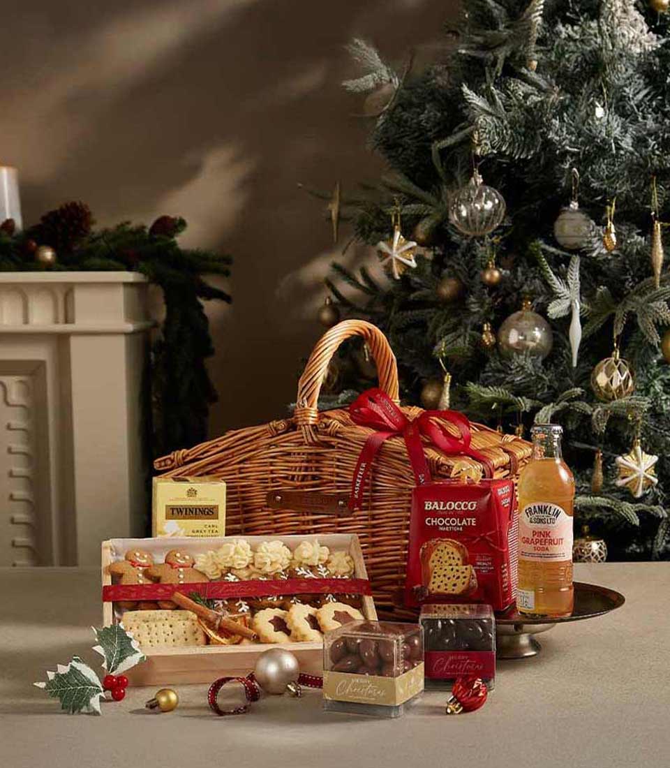 A wicker gift basket filled with Twinings tea, Balocco chocolate, cookies, and Franklin pink grapefruit soda, set in a cozy holiday setting with a decorated Christmas tree.