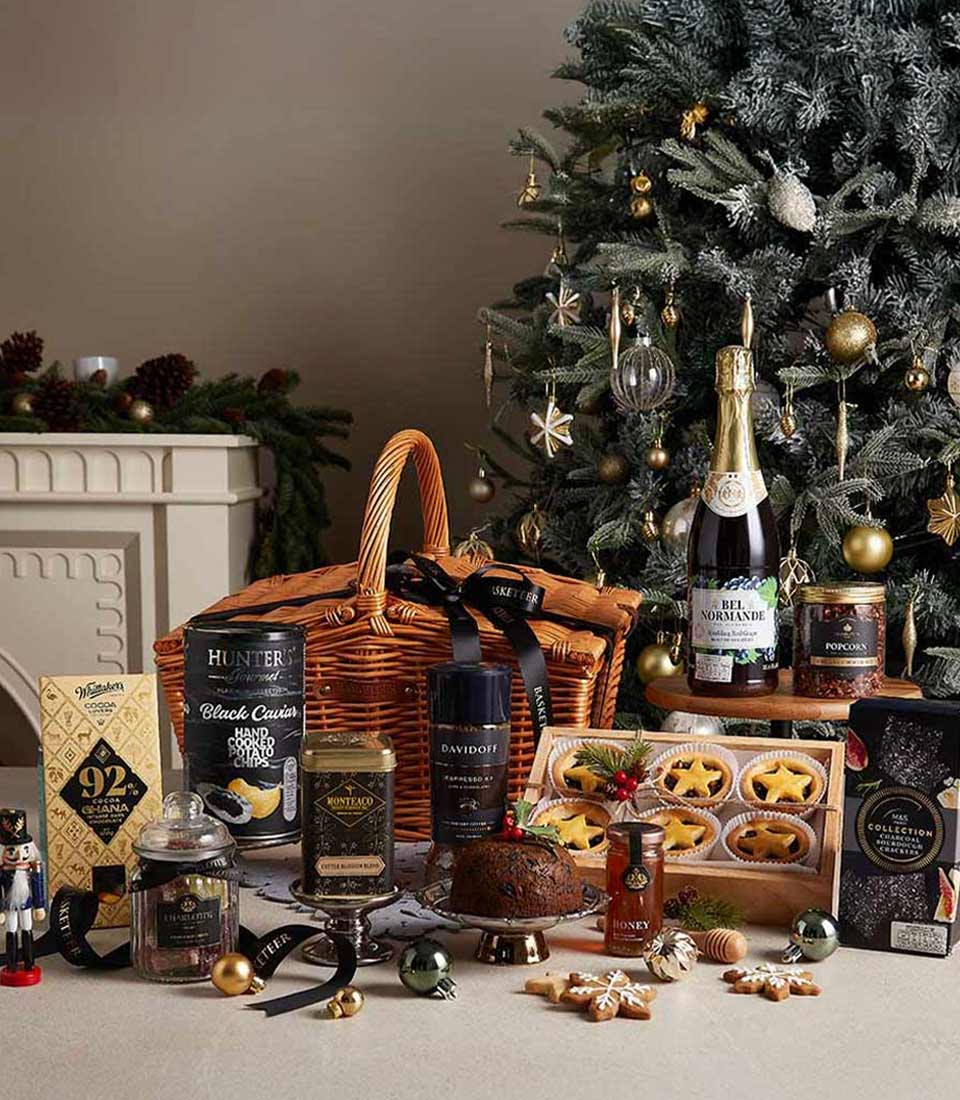 Ultimate gourmet Christmas hamper with luxury coffee, hand-cooked chips, festive treats, and pudding near a Christmas tree.