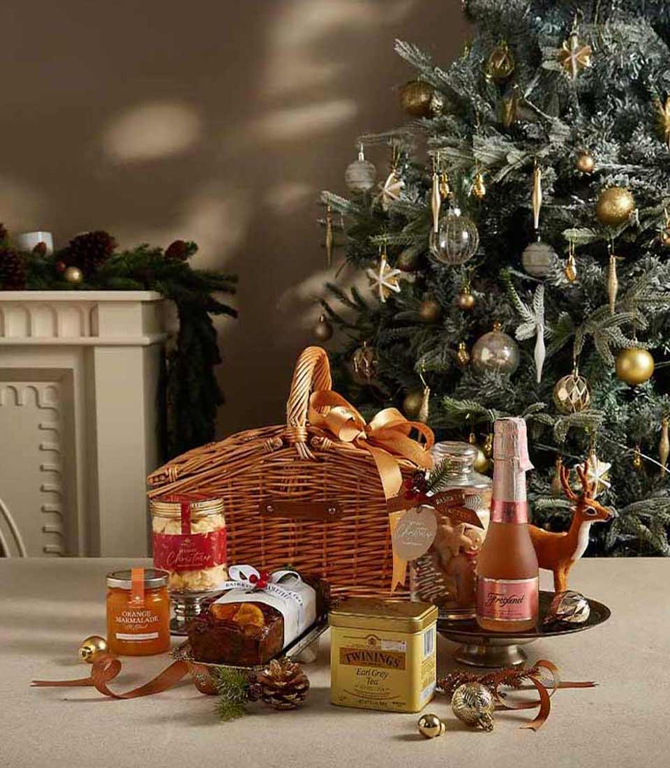 Elegant Christmas hamper with Earl Grey tea, orange marmalade, fruit cake, and Freixenet Rosé, displayed near a beautifully decorated Christmas tree.