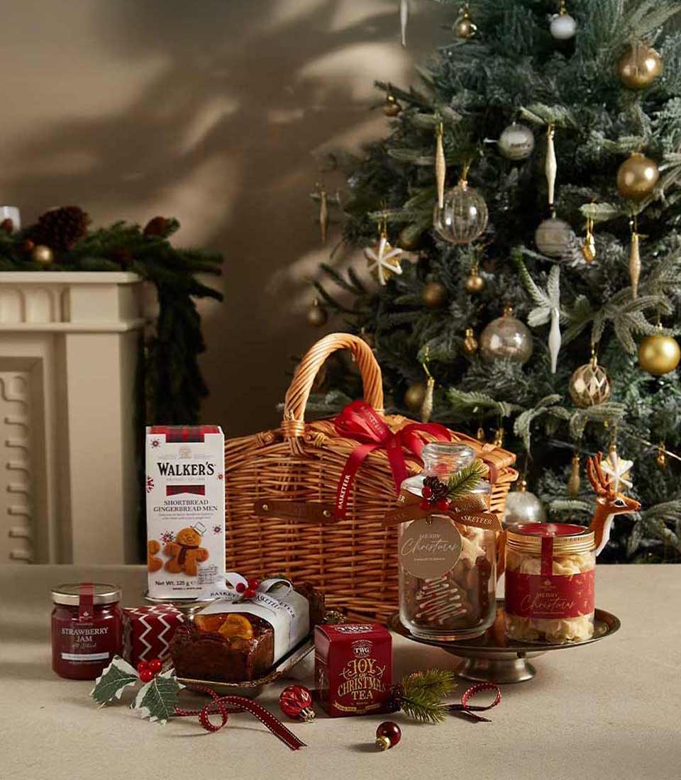 The Christmas Joy Gift Hamper includes a charming wicker basket filled with jars of cookies and jam, chocolate, and a tea canister. Set on a wooden table, it sits against the backdrop of a decorated Christmas tree with festive ornaments and greenery.