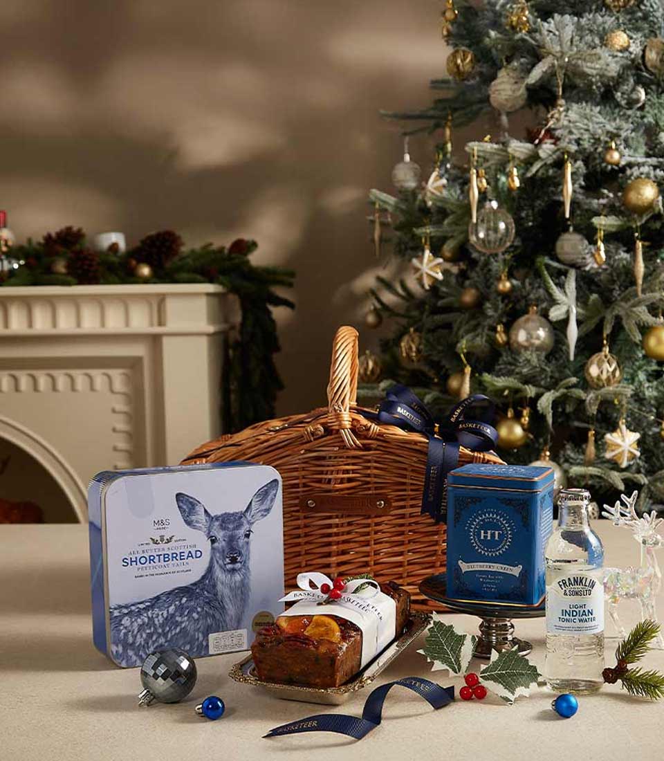 A wicker gift basket with navy ribbon filled with M&S winter-themed shortbread, Harney & Sons tea, Franklin tonic water, Christmas fruitcake, and festive decorations by a Christmas tree.