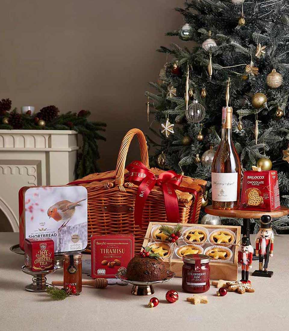 Deluxe Christmas hamper with shortbread, tiramisu, pudding, strawberry jam, and festive tea, beautifully displayed near a Christmas tree.