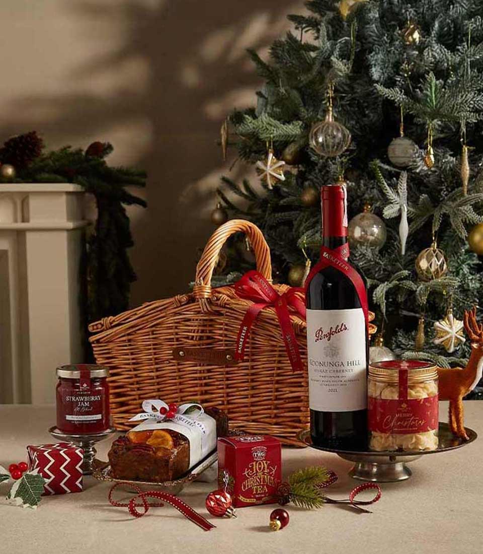 A luxurious Christmas hamper with Penfolds wine, fruit cake, TWG Christmas Tea, strawberry jam, and cookies, set against a festive backdrop with a Christmas tree.