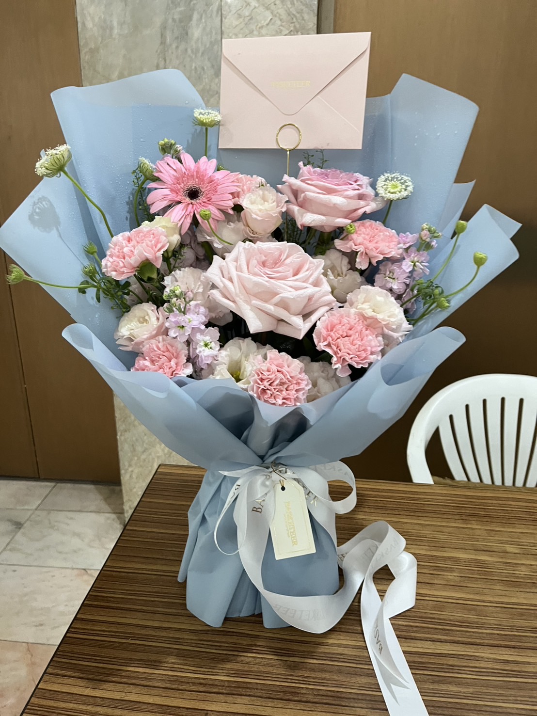 Pastel bouquet with pink roses, gerberas, carnations, and mixed blooms wrapped in soft blue paper with Basketeer ribbon.