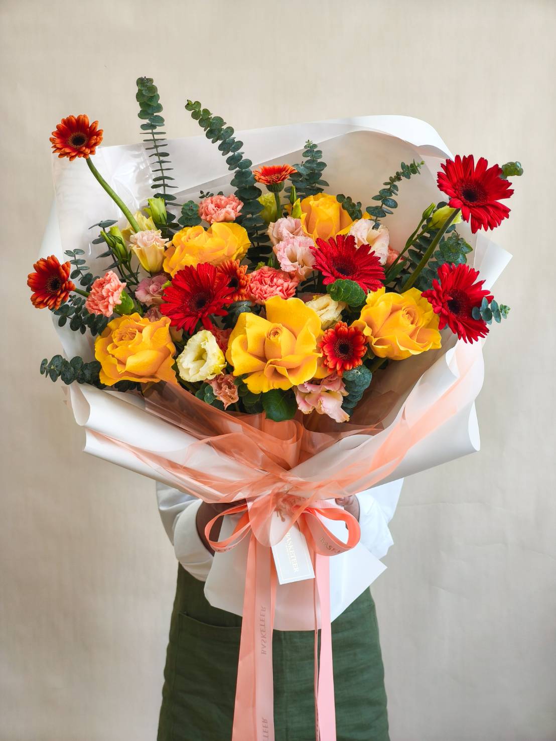 Vibrant bouquet with yellow roses, red gerberas, pink carnations, and eucalyptus, wrapped in white and peach paper with ribbon.