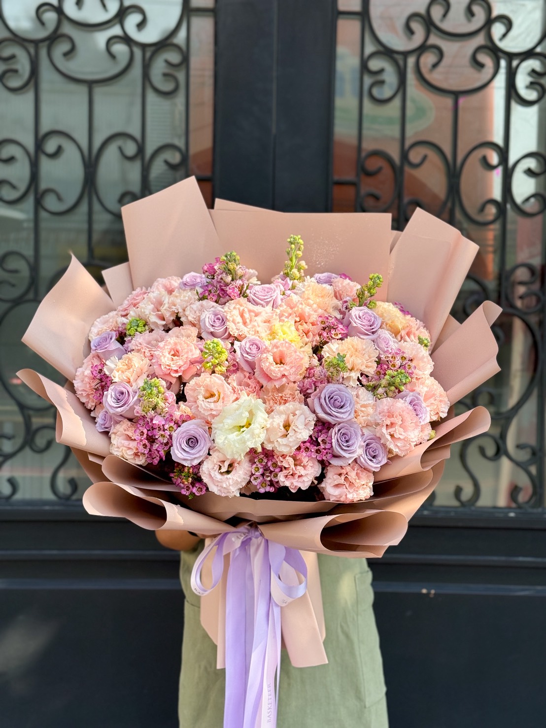 A person holds the Lilac Serenity Luxe Floral Bouquet—pastel pink, purple, and cream blooms wrapped in tan paper with a lavender ribbon—standing before ornate black wrought iron doors.