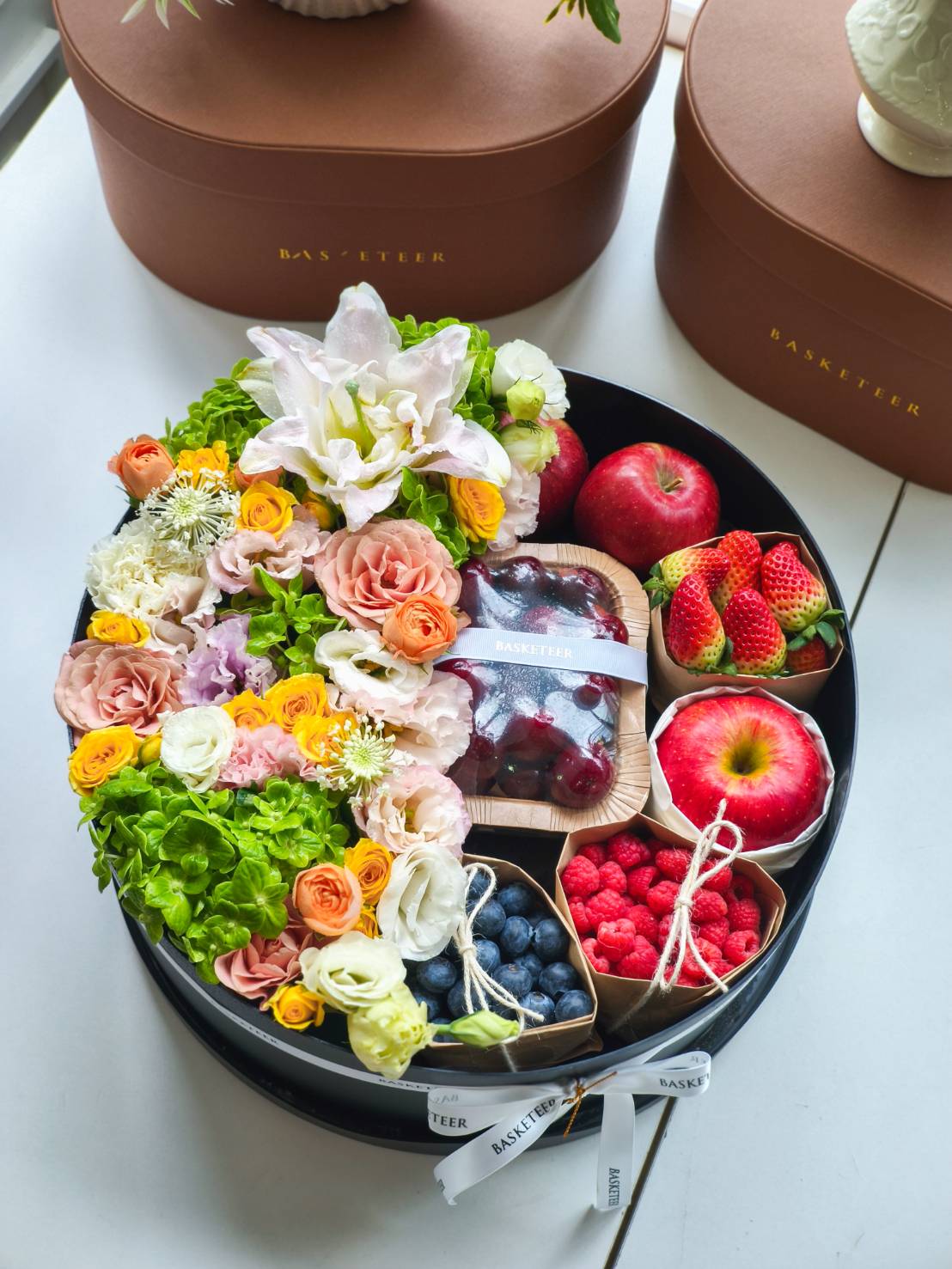 Luxury round black box filled with fresh fruits including apples, strawberries, blueberries, raspberries, cherries, and decorated with colourful flowers