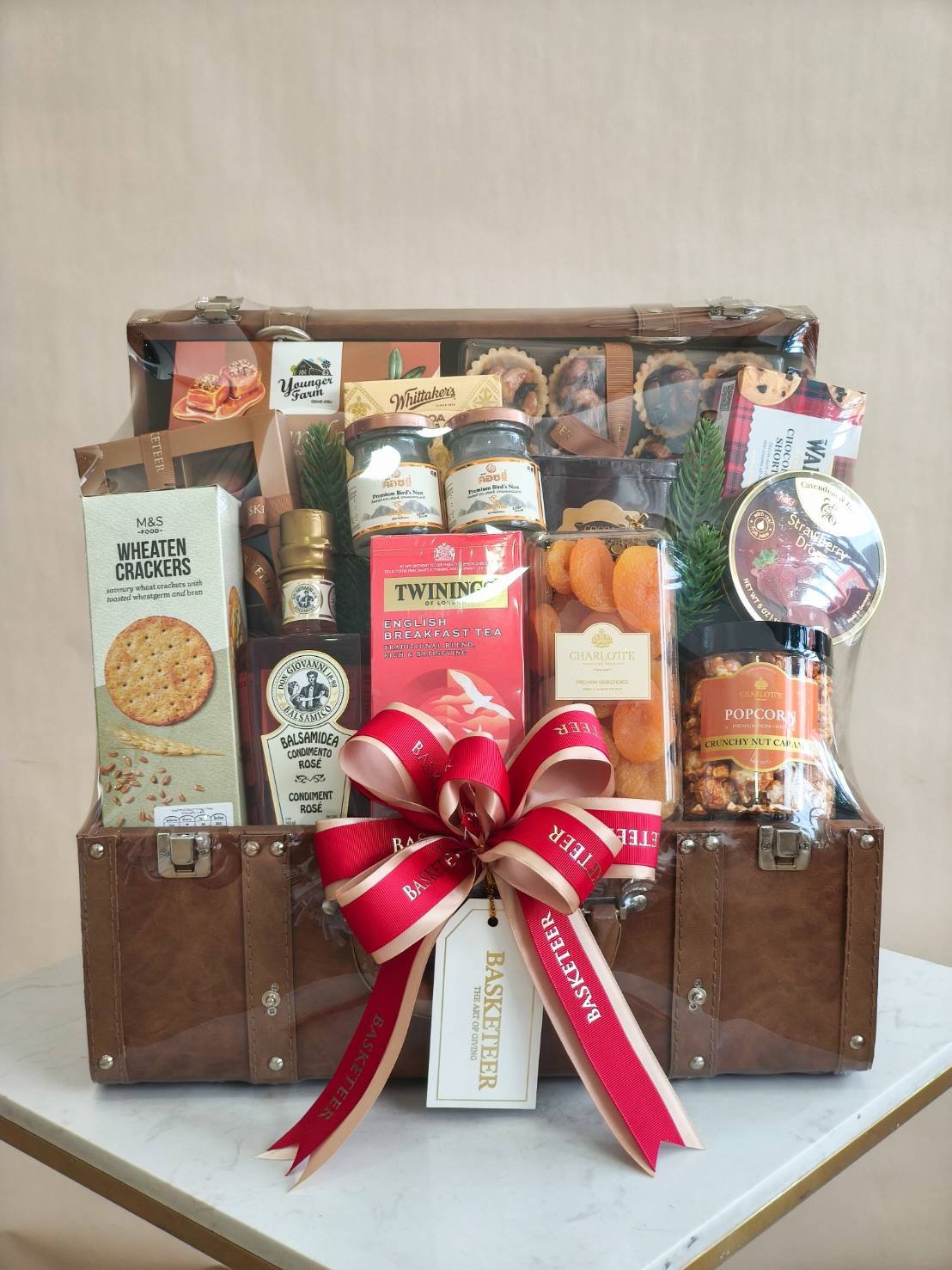 Luxury leather trunk hamper filled with gourmet treats, tea, biscuits, chocolates, balsamic, dried fruits, and popcorn, tied with red ribbon