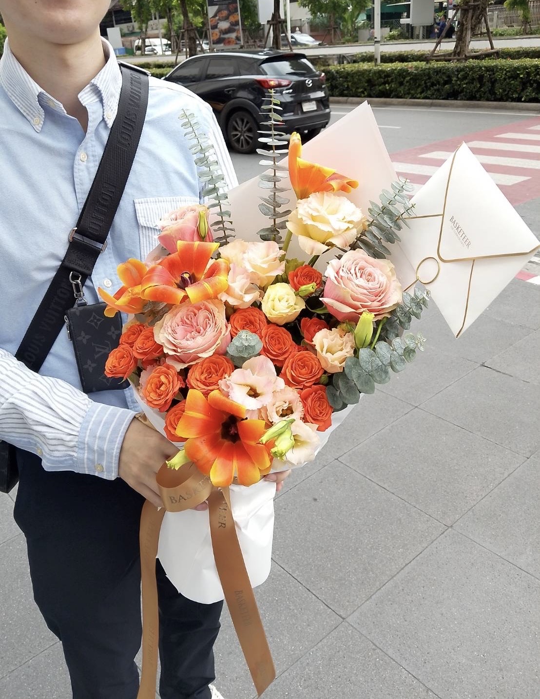 A person in a light blue shirt holds a Vibrant Mixed Flowers Bouquet with orange and peach blooms, greenery, and white wrapping paper, standing on a city pavement near a street with cars in the background.