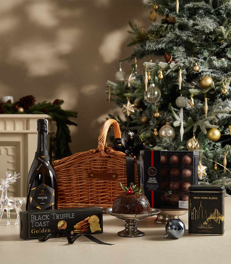 A wicker basket with black ribbons filled with black truffle toast, salted caramel truffles, Christmas pudding, New York Blend tea, and prosecco, set against a festive Christmas tree background.